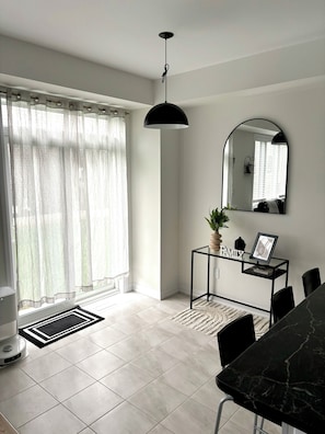 A modern living room with a dining area, a black pendant light, a round mirror, and a glass coffee table with a framed picture.