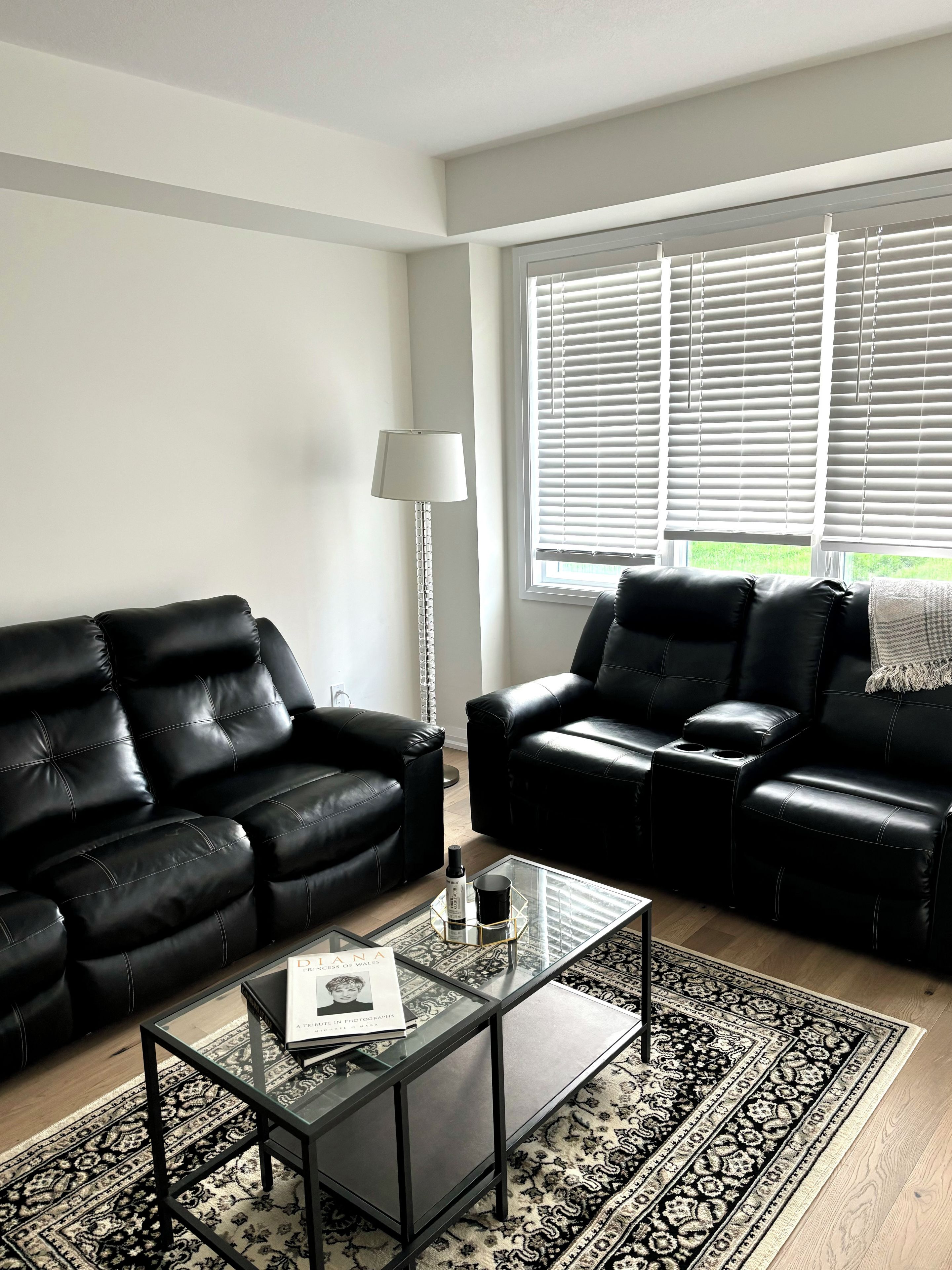 A modern living room with two black leather sofas, a glass coffee table with a book, a floor lamp, and a patterned rug.