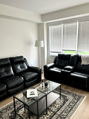 A modern living room with two black leather sofas, a glass coffee table with a book, a floor lamp, and a patterned rug.