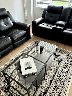 A modern living room with black leather sofas, a glass coffee table with a book and a bottle, and a patterned rug. - The Comfy Couch | Modern 3BR HOME+ Private Jacuzzi (Haldimand)