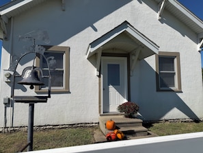 Une maison à l’extérieur blanc, avec un porche à l’avant, une cloche et des citrouilles sur les marches.