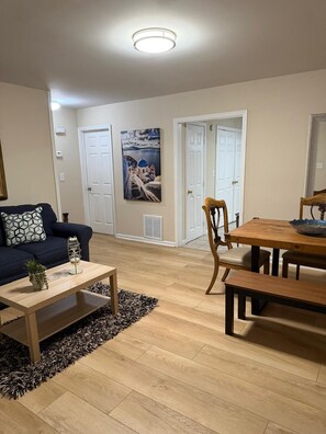 A living room with a sofa, coffee table, and dining table with chairs. There is a painting on the wall and a ceiling light. - The Secret Oasis (Lilburn)