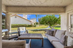 An outdoor patio area with a wooden table, a sofa, and a wicker chair. There is a view of a building and a tree in the background. - Desert Falls Escape Golf, Pools & Mountain Views (Palm Desert)