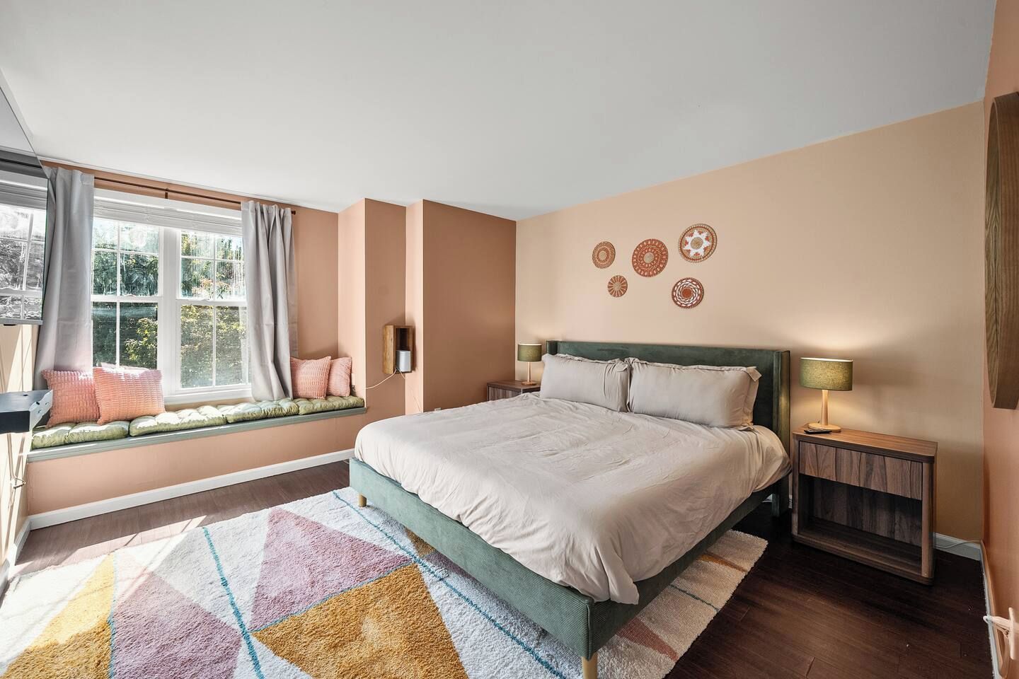 A bedroom with a large bed, a window seat with cushions, a bedside table with a lamp, and decorative wall art.