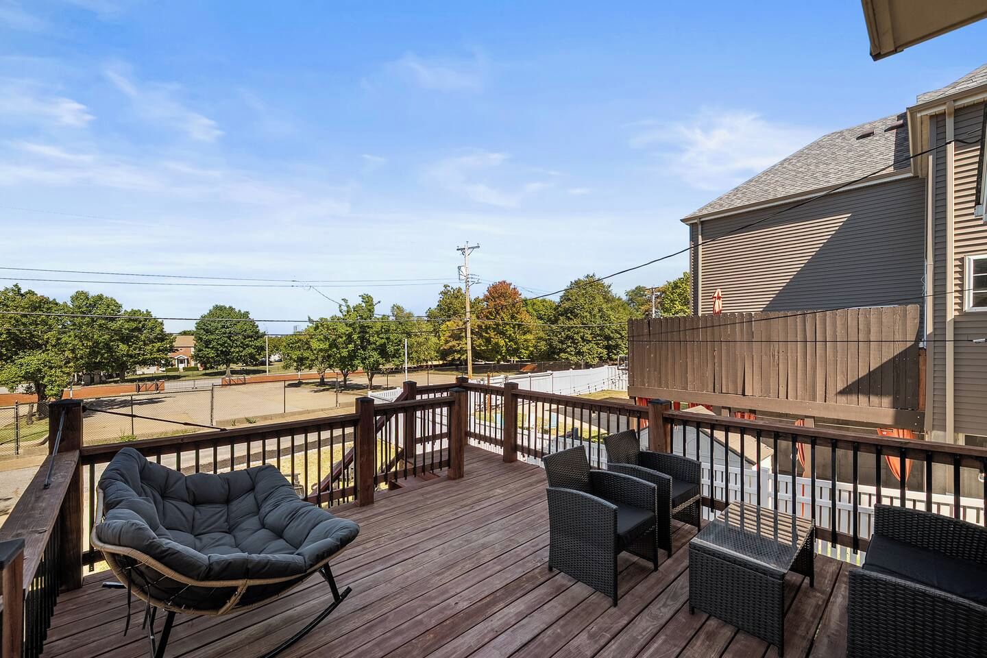 Terraza de madera con mobiliario exterior, que incluye una mecedora y sillas de mimbre, con vistas a un estacionamiento y árboles.