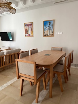 Uma área de refeições com mesa e cadeiras de madeira, televisão na parede e duas pinturas emolduradas.