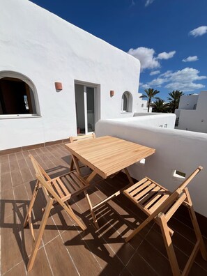 Un edificio blanco con un patio adoquinado, una mesa de madera y dos sillas de madera.