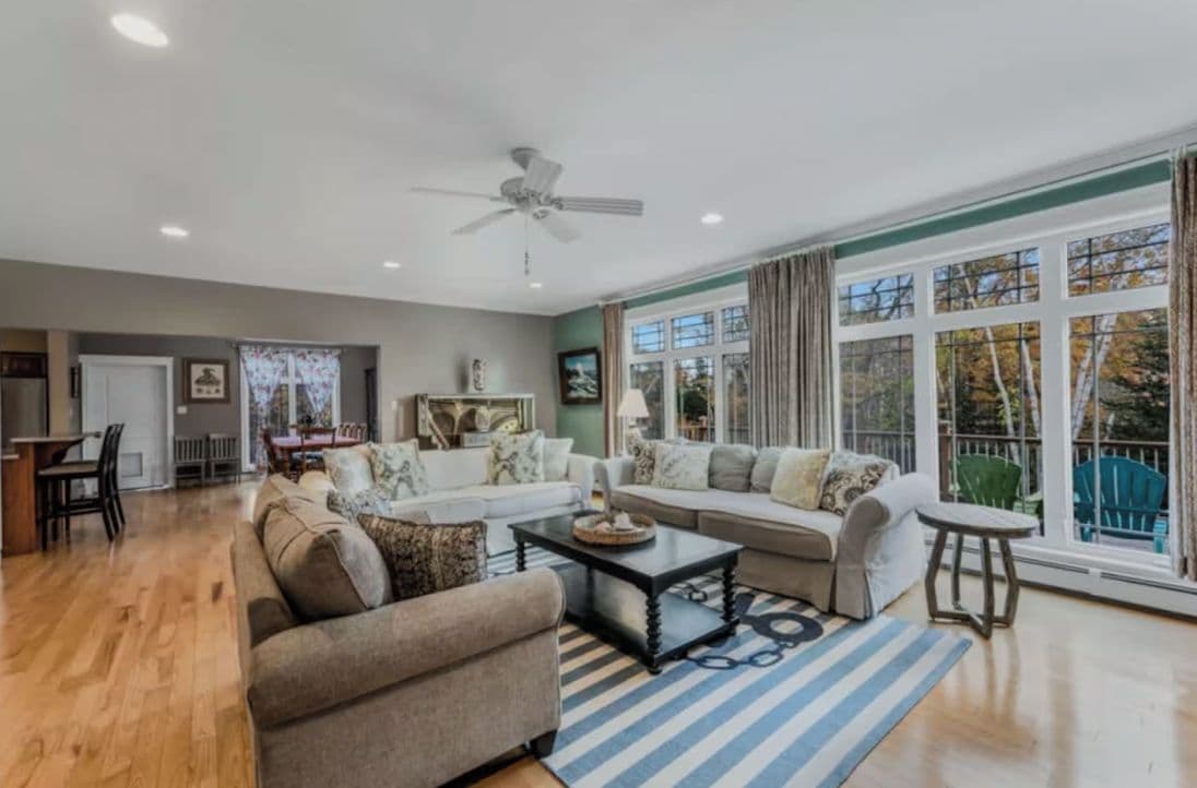 A spacious living room with a large window, a ceiling fan, and a dining area visible in the background.