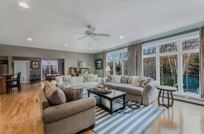 A spacious living room with a large window, a ceiling fan, and a dining area visible in the background. - The Lobster Cottage - Acadia National Park Retreat (Lamoine)