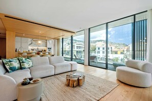 A modern living room with a sofa, armchair, coffee table, and a view of a cityscape through large windows. - Luxury Three-Suite Residence at Savoy Insular (Funchal)