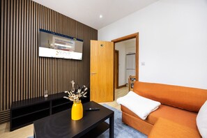 A modern living room with a flat-screen TV mounted on a wooden wall, a sofa, a coffee table, and a door leading to another room. - West Kensington 2BR | 1-Min Tube | Heathrow Direct (Greater London)