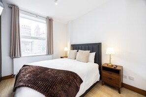 A hotel room with a large bed, two bedside tables with lamps, a window with curtains, and a view of buildings outside. - West Kensington 2BR | 1-Min Tube | Heathrow Direct (Greater London)