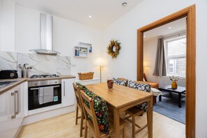 Een compacte keuken met een gasfornuis, oven en witte kastjes. Een eettafel met stoelen staat centraal, en op de achtergrond is een zithoek met een tafel en stoelen te zien.
