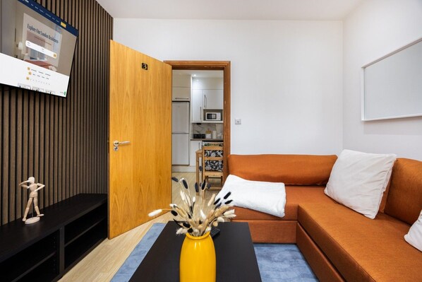 A modern living room with a wooden door, a sofa, a small table with a vase, and a kitchen in the background. - West Kensington 2BR | 1-Min Tube | Heathrow Direct (Greater London)