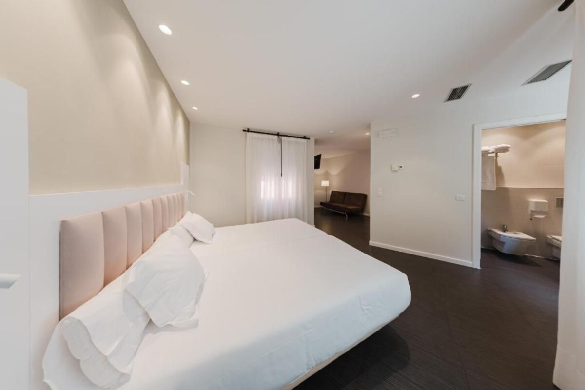 Una camera d'hotel moderna con un letto grande, un bagno con toilette e lavandino, e un'area salotto con una poltrona.