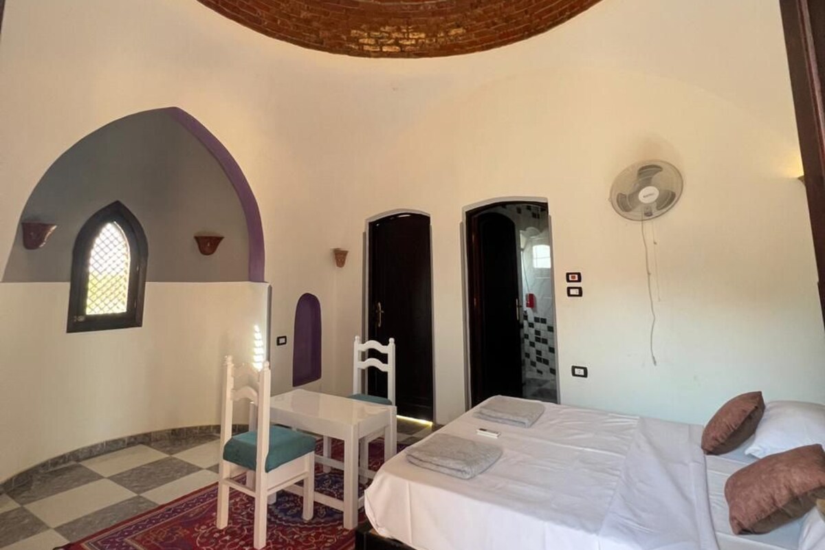 A bedroom with a bed, a table, a chair, a patterned rug, and a unique circular ceiling design.