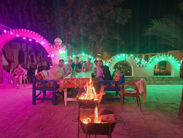 An outdoor gathering with people seated around a fire pit, under illuminated arches, and a decorative sphere on a structure. - Layla EcoResort (Luxor)