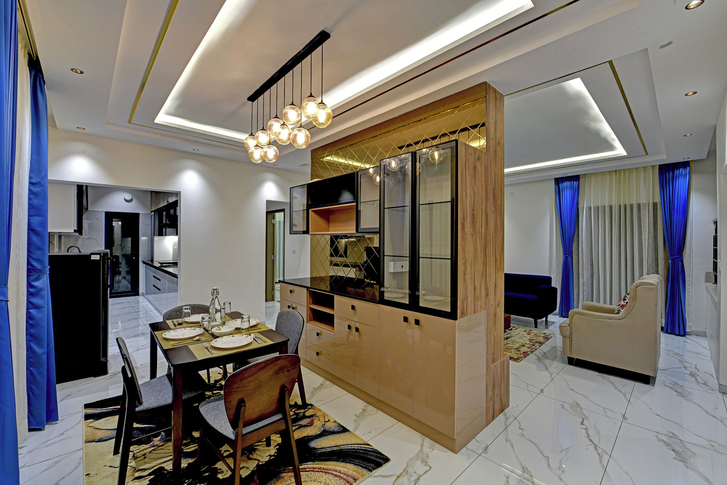 Un espace repas moderne avec un îlot central, une cuisine dotée d’un réfrigérateur intégré et un salon avec un canapé et des rideaux.