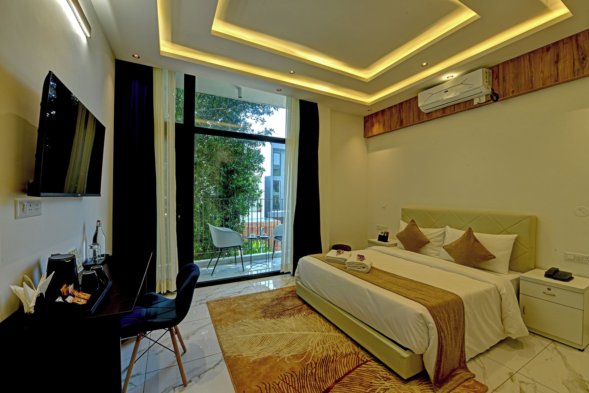Habitación de hotel con cama, mesita de noche y escritorio con silla. Hay un balcón con vistas a zonas verdes.