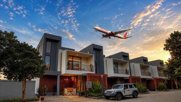 Un moderno edificio residencial de varios pisos con un SUV estacionado, bajo un cielo despejado y un avión volando.