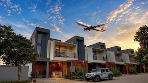 Un moderno edificio residencial de varios pisos con un SUV estacionado, bajo un cielo despejado y un avión volando.