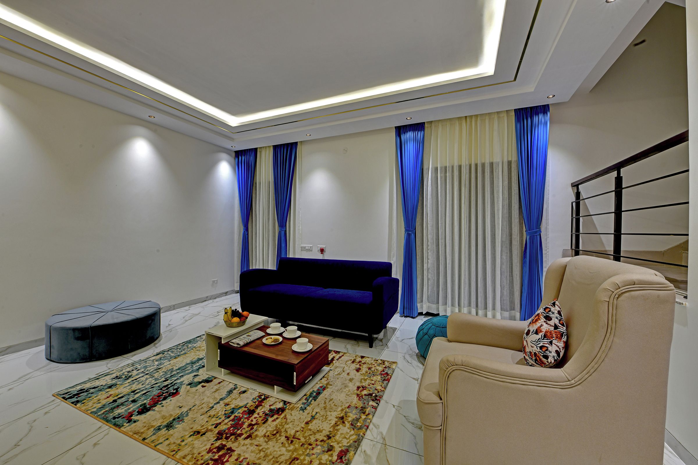 A modern living room with a sofa, a coffee table with fruits, a patterned rug, and a staircase.