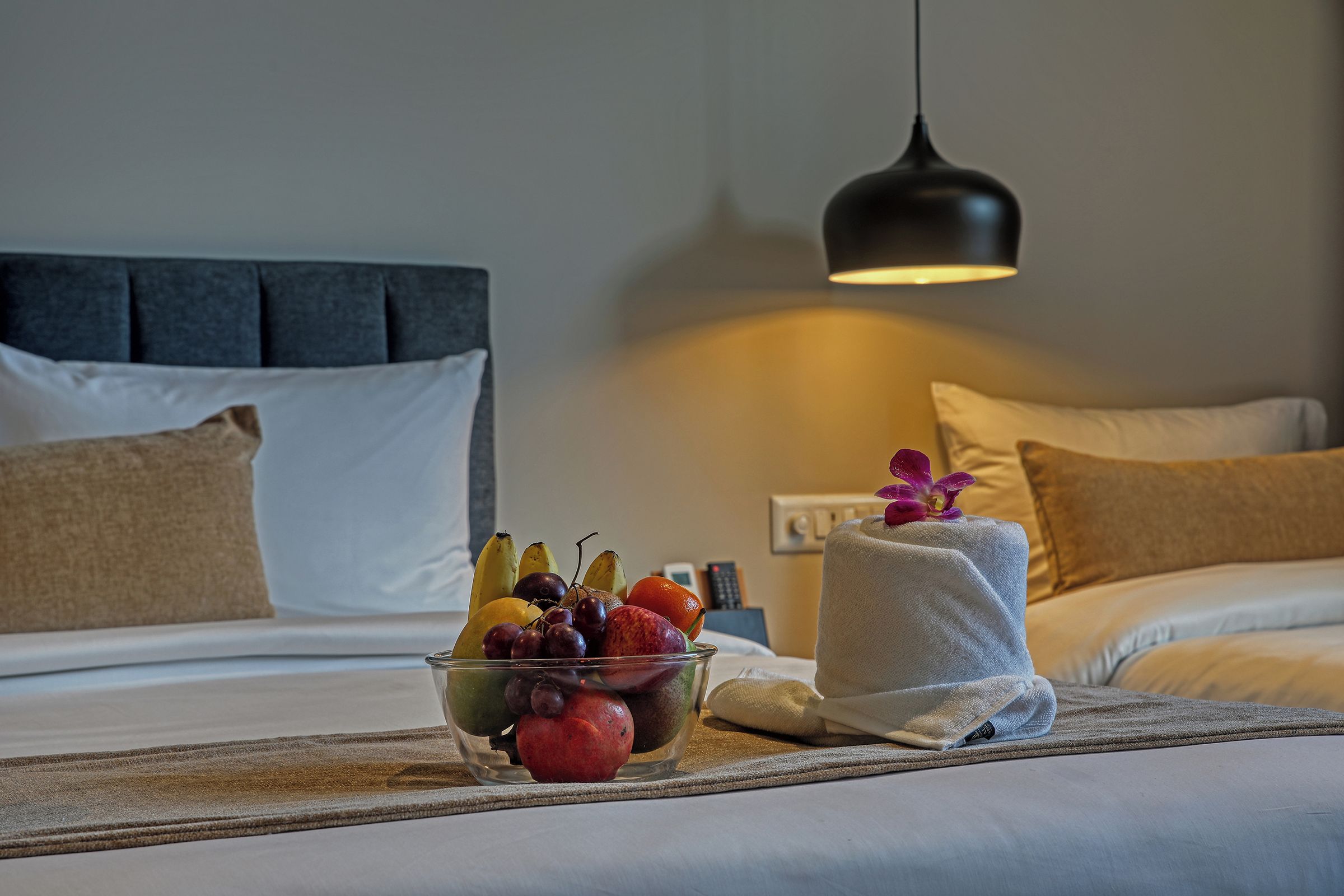 Une chambre d’hôtel avec un lit, une table de chevet sur laquelle se trouve un panier de fruits, une serviette et une lampe suspendue.