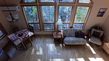 A cozy room with a dining table, a sofa, and a chair, large windows with a view of trees, and a ceiling fan.