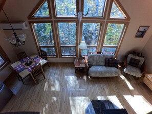 A cozy room with a dining table, a sofa, and a chair, large windows with a view of trees, and a ceiling fan.