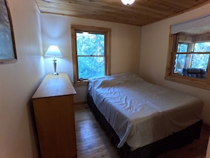 Un dormitorio con una cama individual, una mesita de noche, una lámpara y dos ventanas.