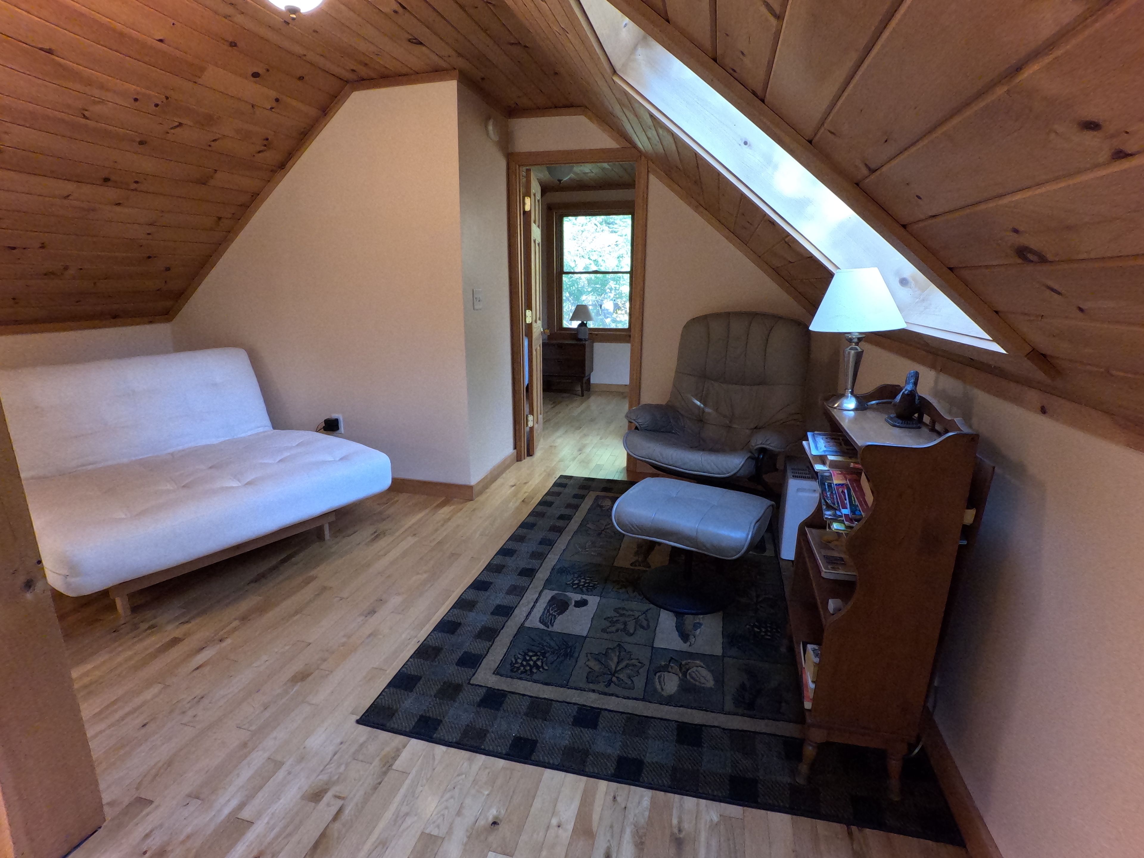 A cozy attic bedroom with a wooden ceiling, a white sofa, a chair, a desk with a lamp, and a patterned rug.