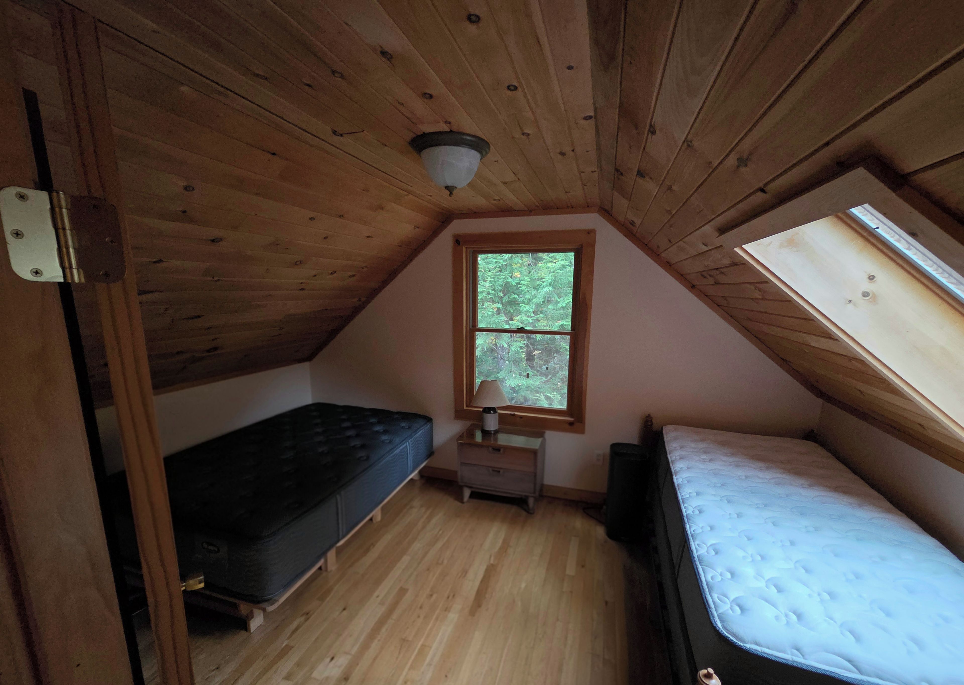 Une petite chambre à coucher de style mansardé, avec un plafond en pente, une fenêtre donnant sur les arbres, une table de chevet avec une lampe et deux lits.