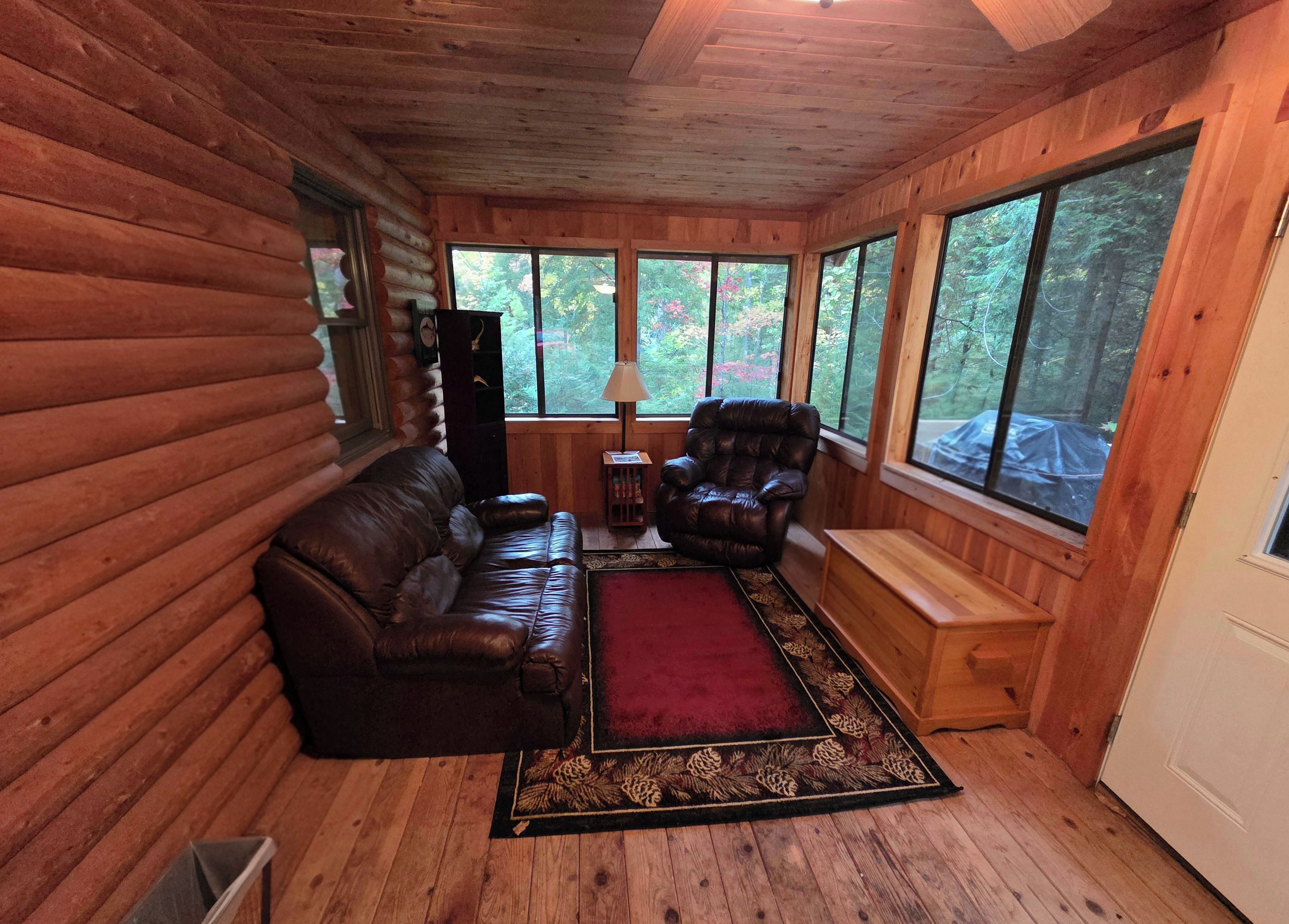 L’intérieur d’une cabane en rondins, avec un canapé en cuir, un fauteuil inclinable, un tapis et de grandes fenêtres donnant sur la forêt.