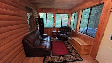 L’intérieur d’une cabane en rondins, avec un canapé en cuir, un fauteuil inclinable, un tapis et de grandes fenêtres donnant sur la forêt.