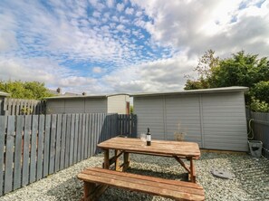 A wooden picnic table with a bottle and glass, surrounded by gravel and a wooden fence. - Holly (Cleator)