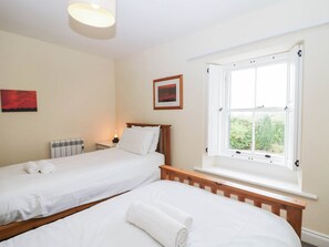 A hotel room with two single beds, a window with a view of greenery, a radiator, and a wall-mounted picture. - Holly (Cleator)