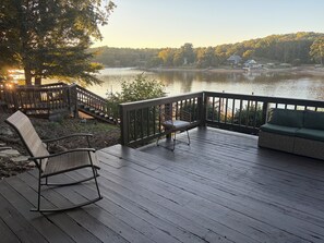 A wooden deck with a sofa and chairs, overlooking a lake with a dock and houses in the background. - The Landing at High Rock Lake-Lakefront-Private Dock-Sleeps 9 (Lexington)