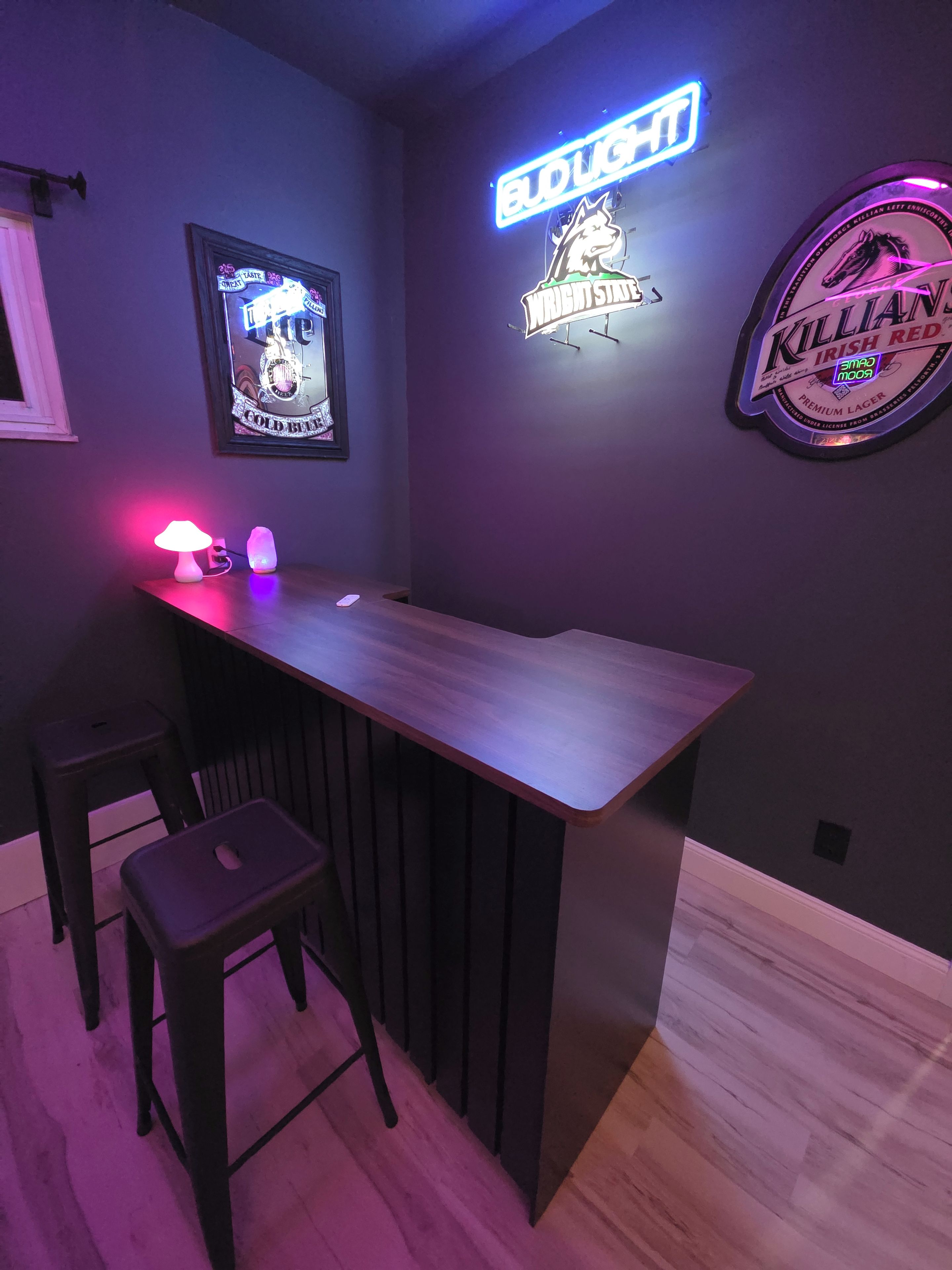 A bar area with a neon sign, a framed poster, and a neon beer logo on the wall.