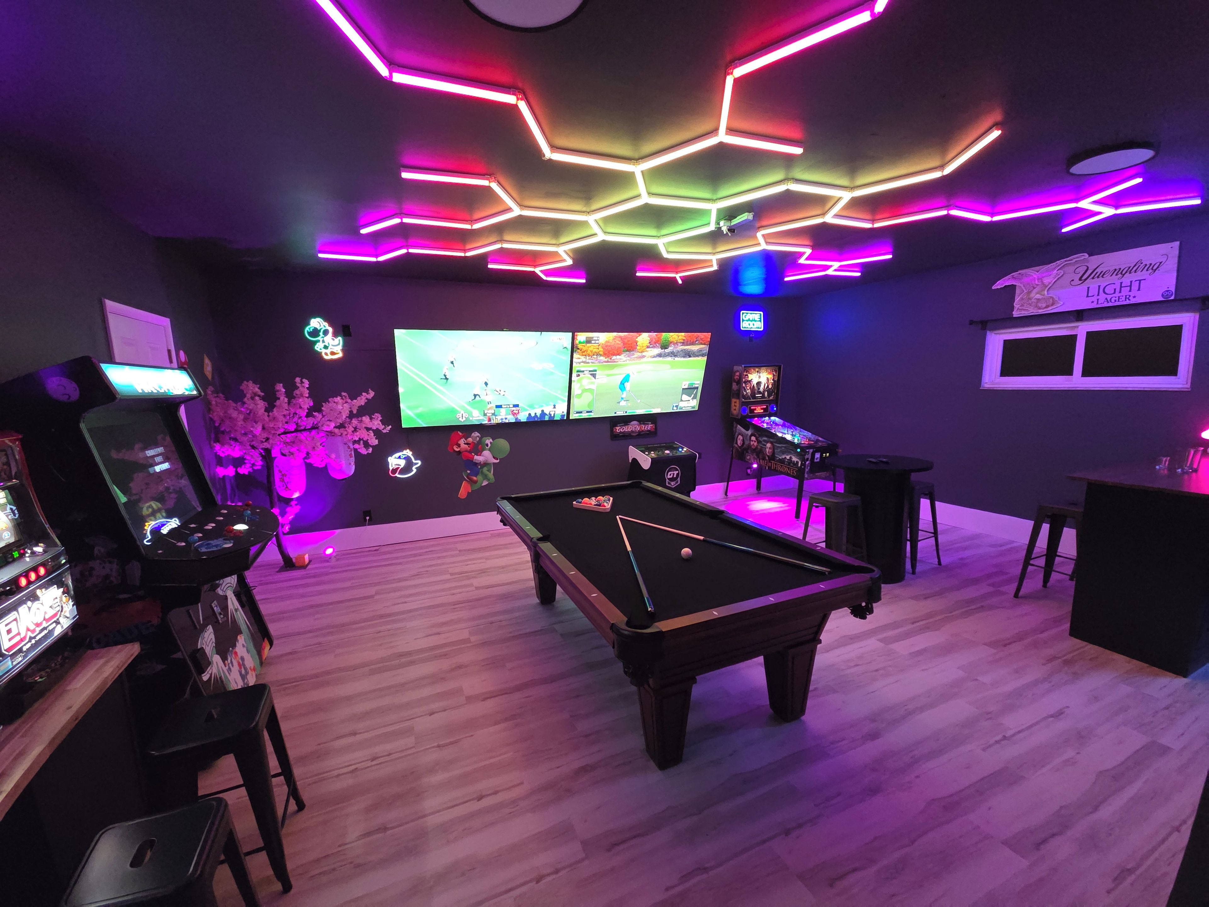 A room with a pool table, gaming machines, and multiple screens displaying sports.