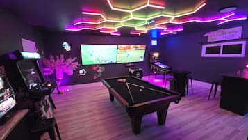 A room with a pool table, gaming machines, and multiple screens displaying sports.