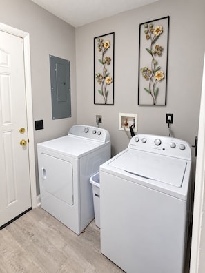 A laundry room with two white front-loading washers and dryers, a gray door, and two framed floral wall art pieces. - Game Room + Hot Tub Near Air Force Museum (Dayton)