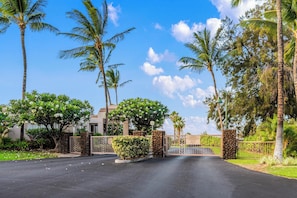A gated entrance with palm trees and a building in the background. - Includes Hilton Pool Pass 2026!  1805 Waikoloa Colony Villas (Waikoloa Beach Resort)