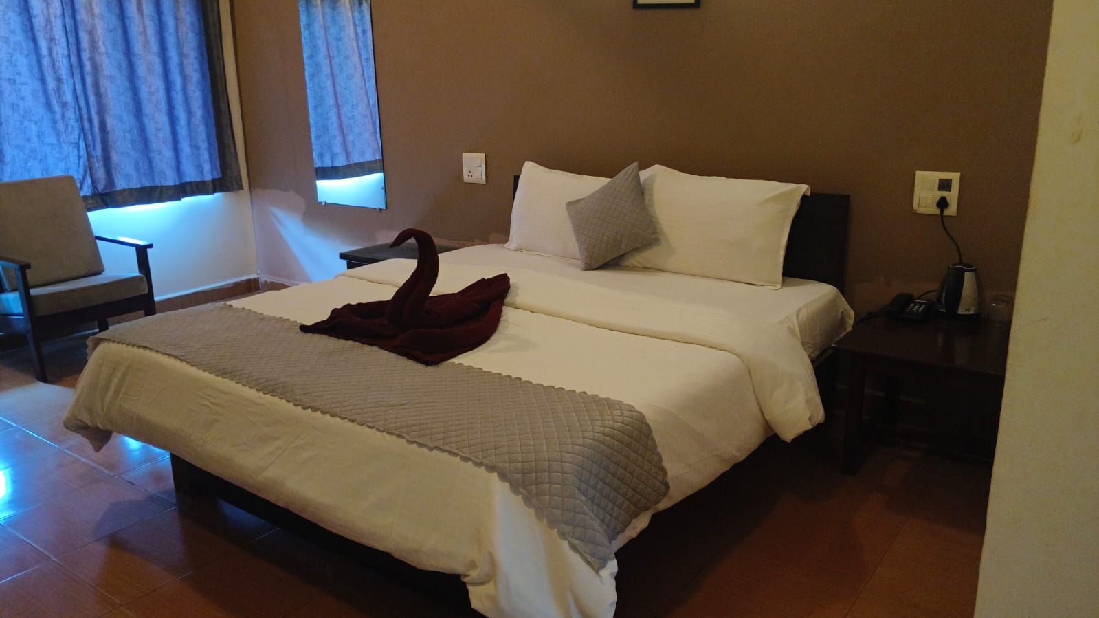 Cama doble con una manta oscura, mesita de noche con un teléfono y una silla en una habitación de hotel.