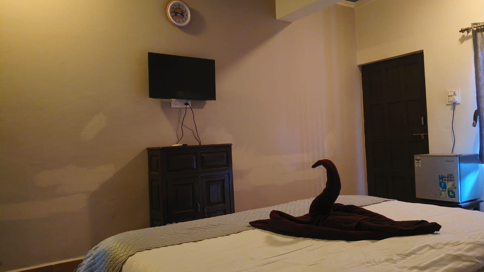A hotel room with a bed, a flat-screen TV mounted on the wall, a dark wooden dresser, and a refrigerator.