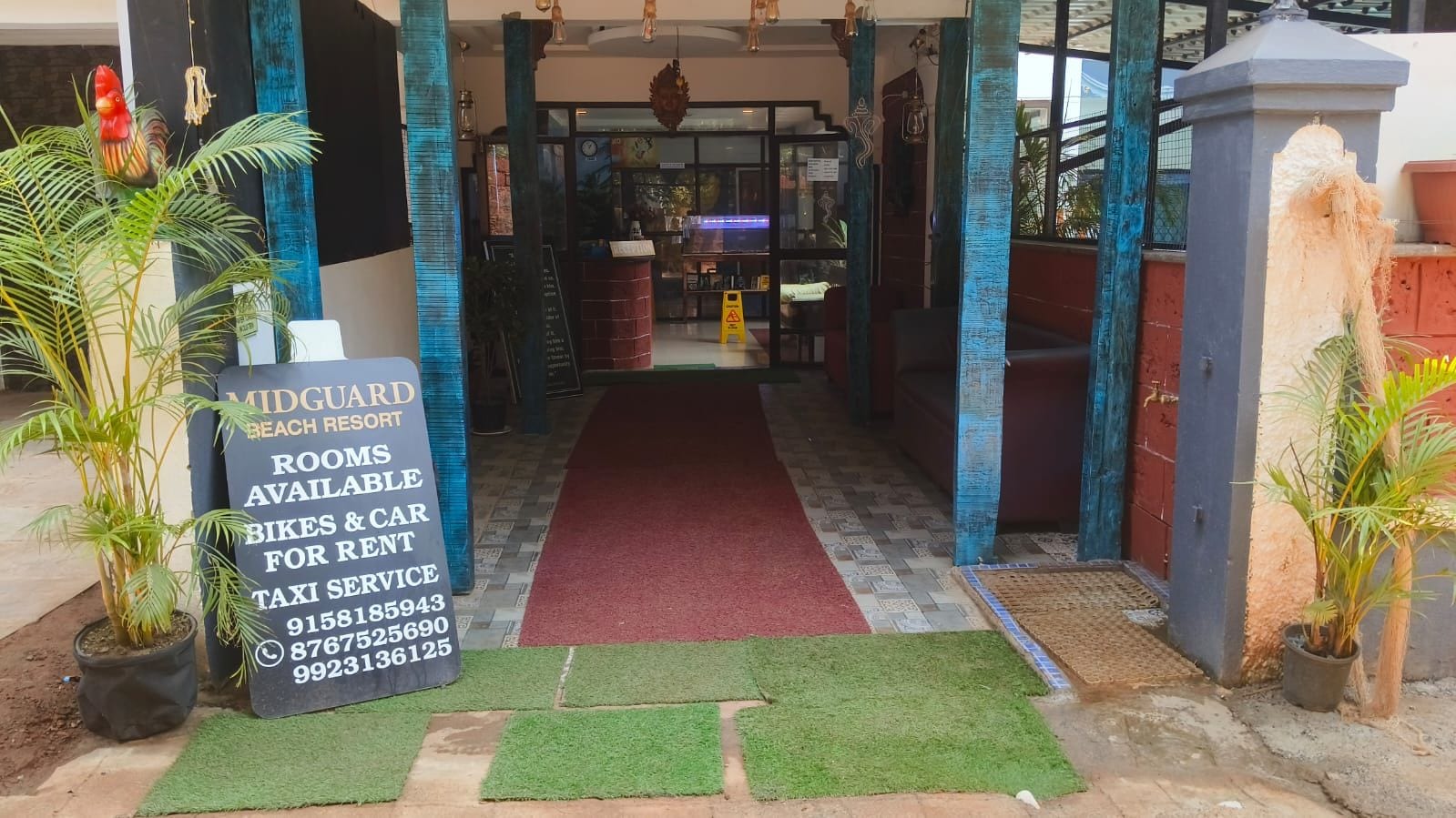 Entrada de hotel com tapete vermelho, plantas em vasos e placa anunciando quartos, aluguel de bicicletas e carros, e serviço de táxi.
