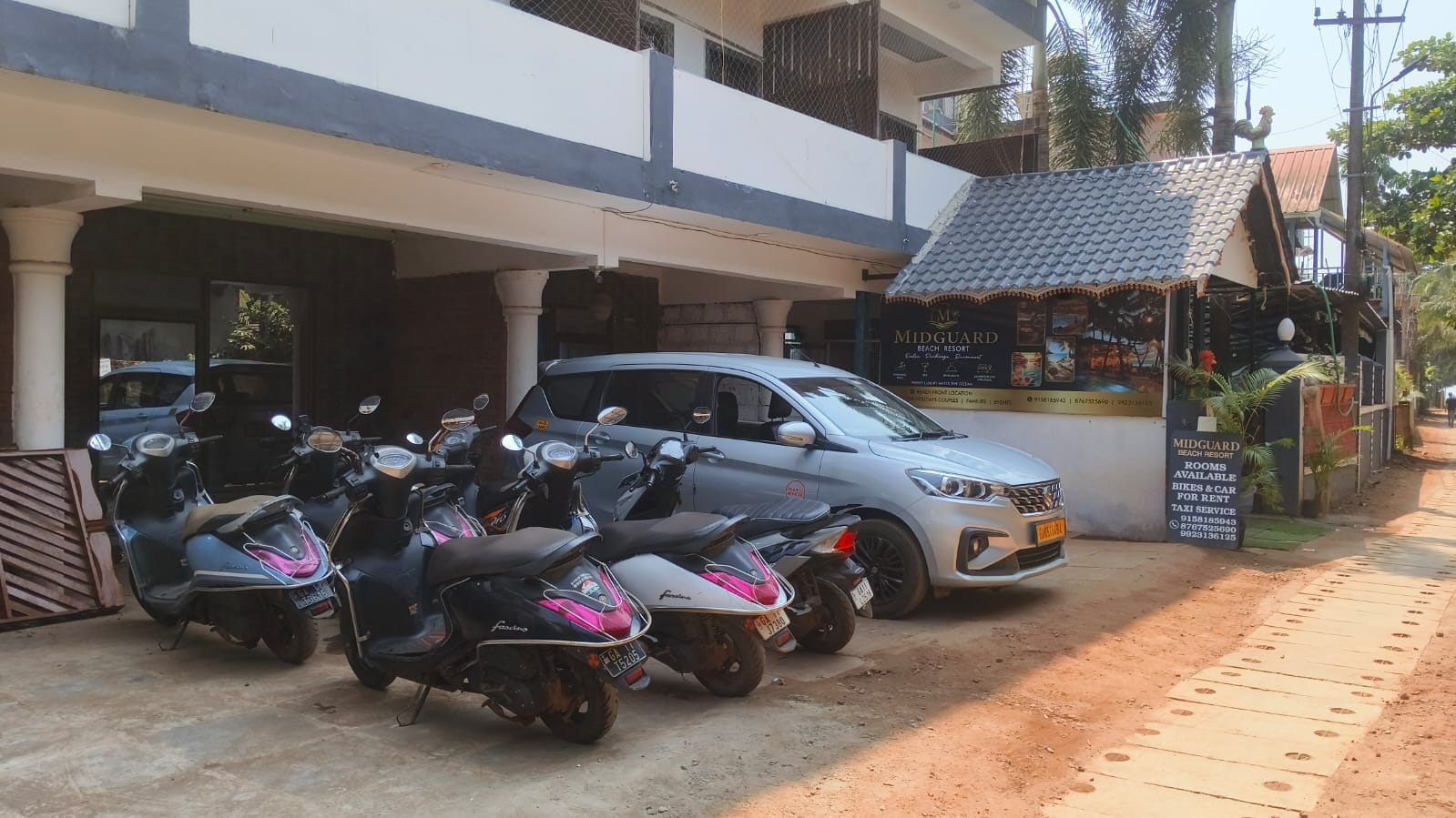 A building with a sign for Midguard Beach Resort, a parked car, and several scooters.