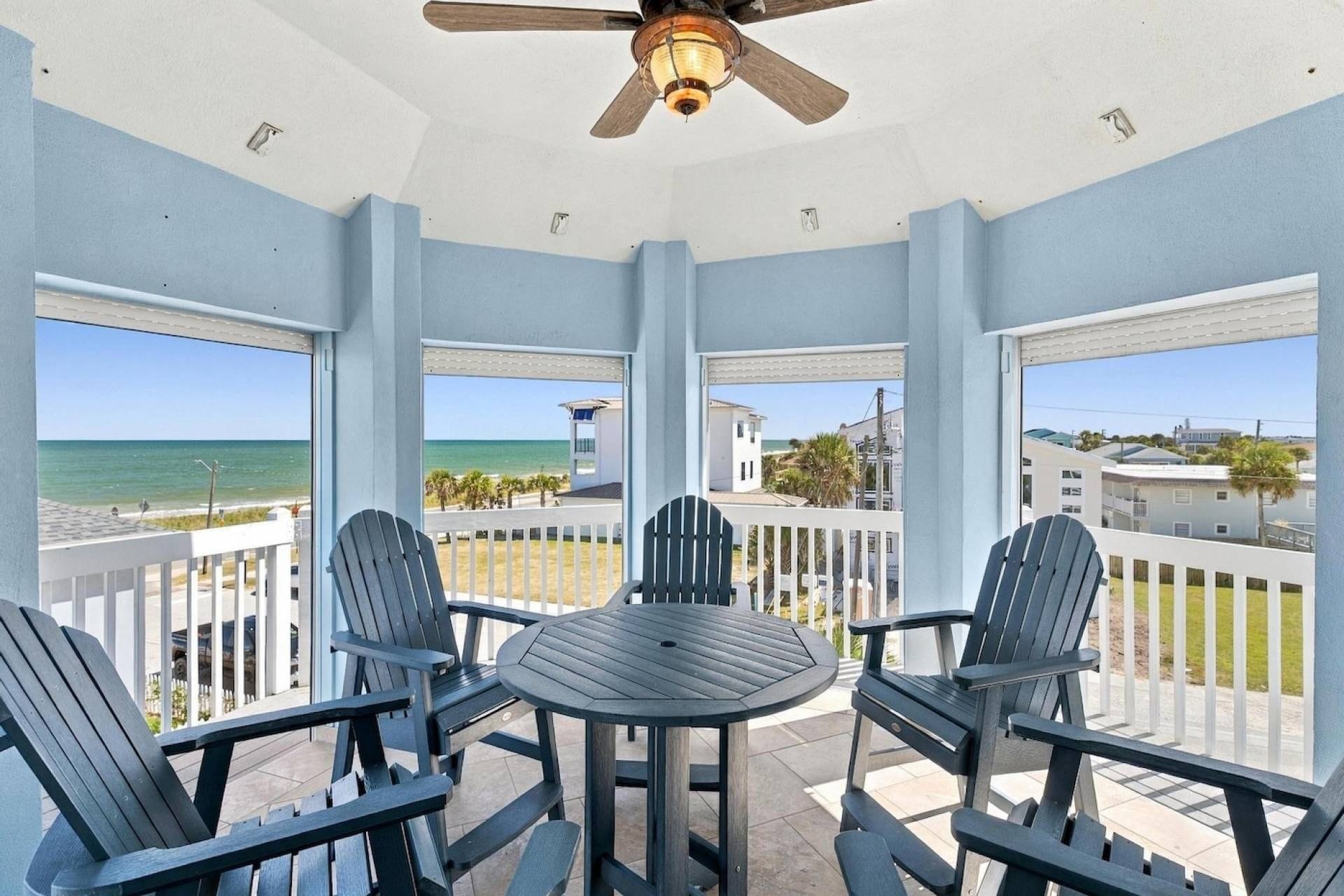 A covered outdoor area with a table and chairs, a ceiling fan, and a view of the beach and ocean.