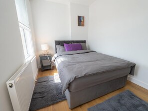 A bedroom with a bed, a nightstand, a lamp, a radiator, and a window. - Stylish 2BR Home with Outdoor Dining/ Social Area (London)