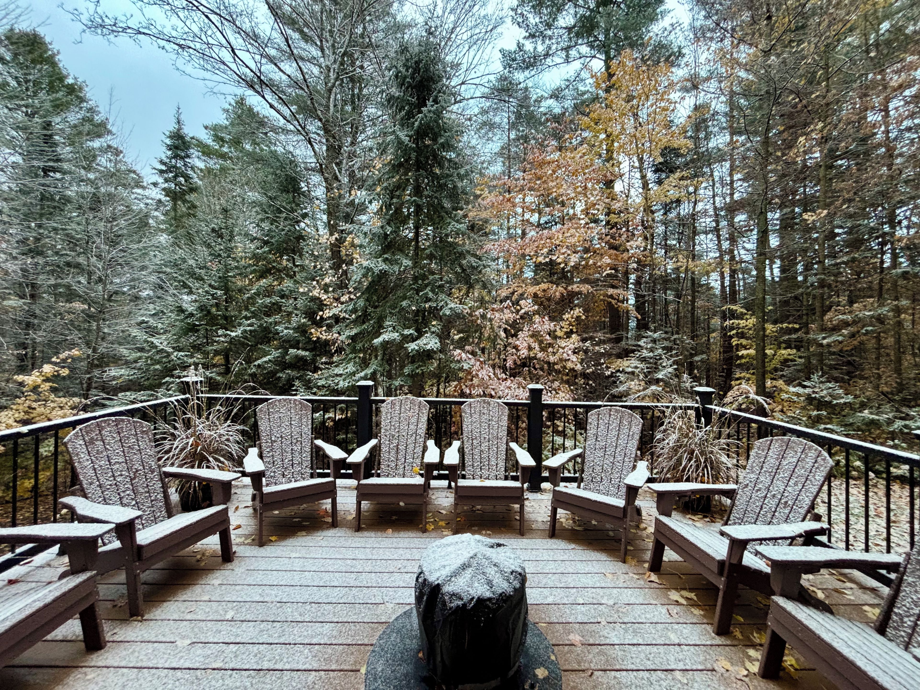A deck with wooden chairs and benches, surrounded by a forest with tall trees.
