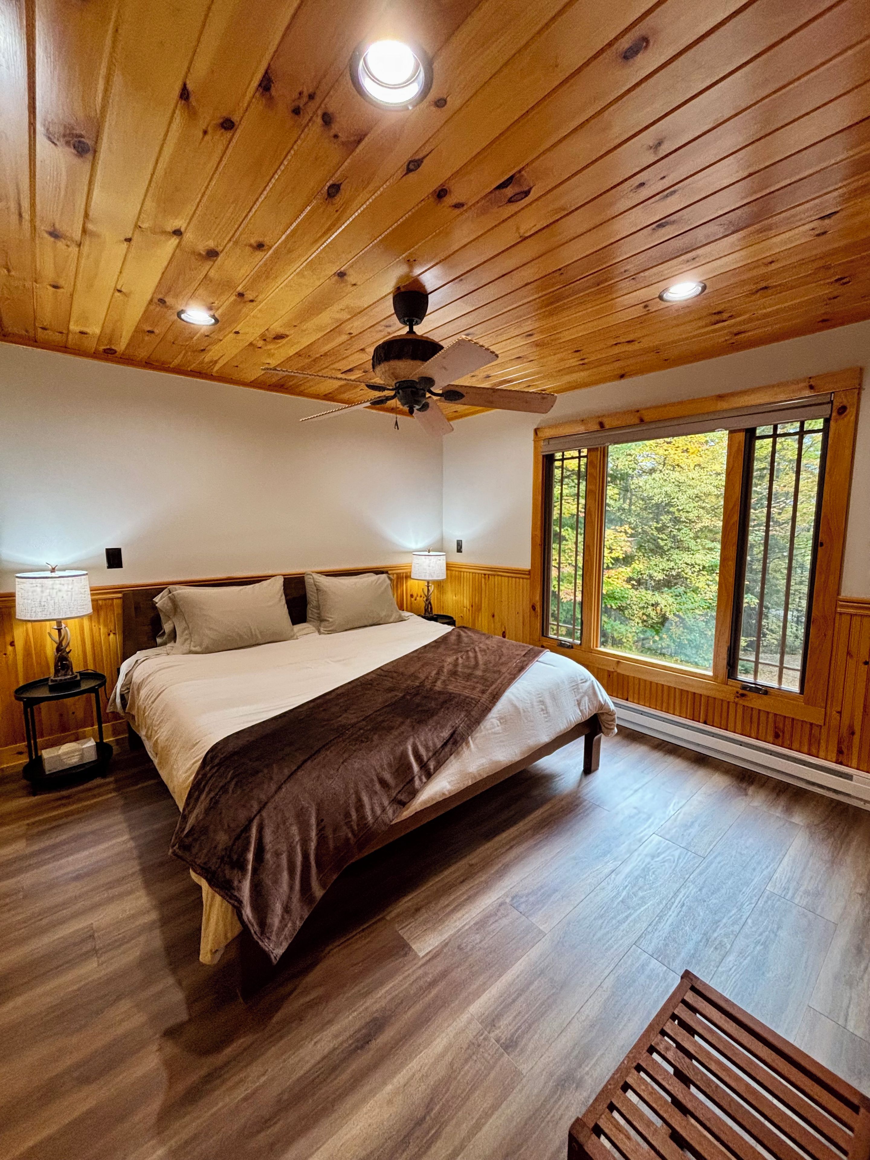 A bedroom with a wooden ceiling, a bed with a brown blanket, a nightstand with a lamp, and a window with a view of trees.